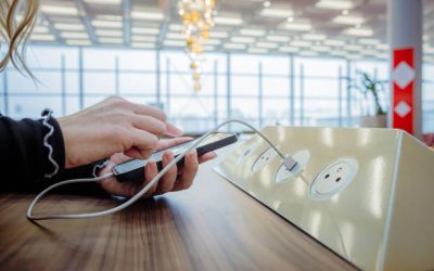 Close-up of a woman with blond hair, wearing a black pullover, using her smartphone while charging it via a white USB cable on a public charging station installed on a wooden table inside an airport terminal, window of the terminal in the background, focus on forefront, horizontal
